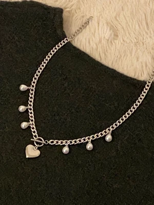 Silverfärgat halsband med pärlor och hjärta - Snyggt silverfärgat halsband med kedja, dekorerat med små vita pärlor och ett hjärtformat hänge. Perfekt accessoar för att lyfta din outfit med en söt och trendig touch. Materialet ser ut att vara metall och pärlorna ger en elegant känsla.