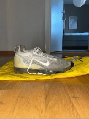 Nike Vapormax flyknit - Nike Air Vapormax sneakers i vitt med transparent sula och stickad ovandel. Skorna har en sportig design med tydlig Swoosh-logga på sidan och vita skosnören. Ovandelen är i mesh-liknande material för bra andningsförmåga och komfort.