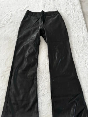 Svarta skinnbyxor med vida ben - Snygga svarta skinnbyxor med hög midja och vida ben. Byxorna har knapp och dragkedja framtill samt bälteshällor. Perfekta för en trendig och edgy look. Längden: Short 