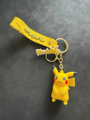 Nyckelring med Pikachu och Pokémon-logga - Säljer en gul nyckelring med Pikachu-figur i mjukt gummi och tillhörande Pokémon-logga. Nyckelringen har även en tag med texten 'Pikachu' och en gul rem. Perfekt accessoar för Pokémon-fans!