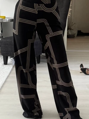 Svarta vida leggings med grafiskt mönster - Säljer ett par svarta leggings med vida ben och ett coolt grafiskt mönster i beige. De har hög midja och är tillverkade i ett mjukt, stretchigt material som sitter bekvämt. Perfekta för dig som vill sticka ut med en unik stil.