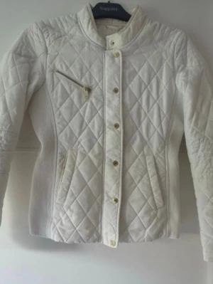 Vit quiltad jacka från Lindex - Snygg vit quiltad jacka från Lindex med ståkrage, guldfärgade knappar och dragkedja på bröstet. Jackan har två fickor fram och ribbade partier på sidorna för extra komfort. Perfekt för dig som gillar stilrena och moderna plagg. Lite nopprig på en ställe, men inte så synligt.