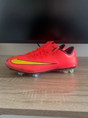 Nike Mercurial Vapor 10 Elite - Säljer ett par Nike Mercurial fotbollsskor i en grymt snygg röd färg med gula och svarta detaljer. Skorna har en låg profil, snörning och plastdobbar som ger bra grepp på planen. Perfekta för dig som vill ha snabbhet och kontroll på matchen.