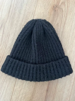 Svart ribbstickad mössa - Säljer en svart ribbstickad mössa med uppvikt kant. Mössan har en klassisk form och är tillverkad i ett mjukt, värmande material som passar perfekt för kyliga dagar. Enkel och stilren design som funkar till det mesta.