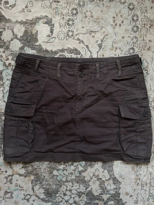 Gröngrå cargokjol med fickor från Brandy - Säljer en gröngrå cargokjol med stora sidofickor och bälteshällor. Kjolen har knapp- och dragkedjestängning framtill och är i en kort modell. Perfekt för en avslappnad och cool stil. Har några jättesmå vita fläckar men syns inte när man har på sig den. Onesize men sitter som S