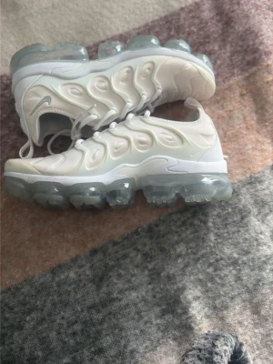 Nike Air Vapormax Plus vita sneakers - Nike Air Vapormax Plus sneakers i vitt med genomskinlig sula och futuristisk design. Ovandelen är i syntetmaterial med vågiga detaljer och klassisk Swoosh-logga på sidan. Skorna har snörning och en chunky, transparent bubbelsula som ger en cool vibe.
