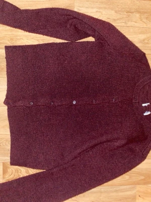 Vinröd kofta med knappar - Mysig vinröd kofta med långa ärmar och knäppning framtill. Enkel och stilren design, perfekt att slänga på sig över en t-shirt. Stickad i mjukt material och har rund halsringning.