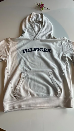 Vit hoodie från Tommy Hilfiger - Vit hoodie från Tommy Hilfiger med mörkblått HILFIGER-tryck på bröstet. Klassisk känguruficka framtill, ribbade muddar och huva. Perfekt för en avslappnad och stilren look.🙌🏼