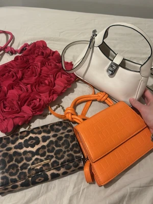 Fyra trendiga handväskor i olika stilar - Fyra handväskor: en vit axelremsväska med silverdetaljer, en orange väska med krokopräglat mönster och knutade axelband, en leopardmönstrad clutch och en rosa väska dekorerad med stora tygrosor. Perfekta för att lyfta din outfit med färg och mönster.