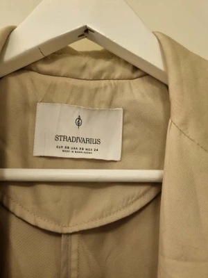 Beige trenchcoat från Stradivarius - Stilren beige trenchcoat från Stradivarius med klassisk krage, knappar och två stora fickor framtill. Lång modell med avslappnad passform och snygga detaljer vid ärmsluten. Perfekt för lager-på-lager och en cool look.