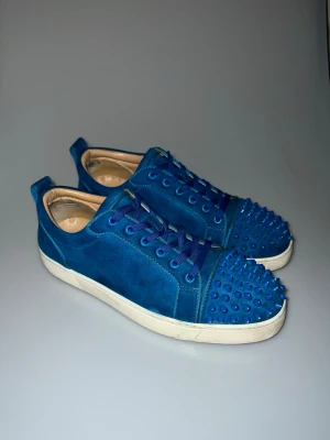 Blåa Christian Louboutin med nitar - 6 nitar är av därmed priset! Unika blå sneakers med mocka material och matchande blå skosnören. Tåpartiet är täckt av coola, rundade nitar som ger en edgy vibe. Skorna har vit röd sula med trycket Louboutin!  och lågt skaft för en clean och modern look. Perfekta för dig som vill sticka ut.