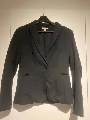 Svart klassisk kavaj med enkel knapp - Stilren svart kavaj med figurnära passform och en enkel knapp framtill. Kavajen har långa ärmar, två fickor och klassisk krage. Perfekt för en snygg och clean look till vardags eller mer uppklätt. Materialet ser ut att vara en blandning av polyester och viskos.