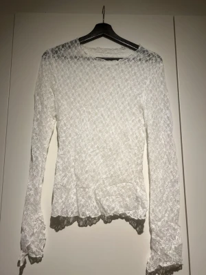 Vit spetstopp med lång ärm - Superfin vit topp i spets med långärmad design och lätt figursydd passform. Toppen har ett romantiskt mönster och volangdetaljer vid ärmslut och nederkant. Perfekt att styla med jeans eller kjol för en trendig look.