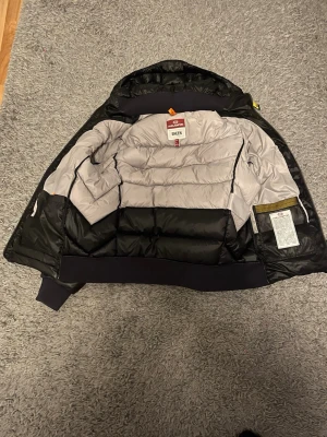 Parajumpers Pharell dunjacka svart - Snygg dunjacka från Parajumpers, modell Pharell i svart med grå insida. Jackan har quiltad design, ribbade muddar och klassisk orange detalj vid nacken. Perfekt för kalla vinterdagar och riktigt varm tack vare dunfyllningen.