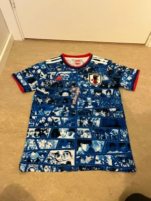 Japan anime fotbollströja blå Adidas - Unik blå Japan fotbollströja från Adidas med anime-motiv över hela plagget. Röd krage och ärmslut, officiellt Japan-märke på bröstet och röd Adidas-logga. På ryggen står det 'ISHOWSPEED' och siffran 7. Perfekt för dig som älskar både fotboll och anime.