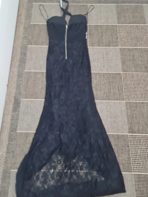 Svart spetsklänning från Isoka - Elegant svart långklänning från Isoka med spetsdetaljer och smala axelband. Klänningen har en markerad dragkedja framtill och en lätt transparent nederdel. Perfekt för dig som vill sticka ut med en sofistikerad look.