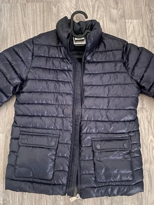 Mörkblå tunn Dunn jacka - Snygg mörkblå dunjacka med quiltad design och hög krage. Jackan har två stora fickor framtill med lock och tryckknappar samt dragkedja. Perfekt för vår dagar och ger en stilren look.