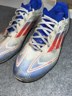 Adidas f50 vita  - Säljer ett par Adidas F50  med vit bas, röda och blå detaljer samt blå plös. Skorna har klassiska snörningar och de ikoniska tre ränderna på sidan. Ovandelen är tillverkad i syntetmaterial för lätt vikt och bra bollkontroll. Perfekta för dig som vill sticka ut på planen.
