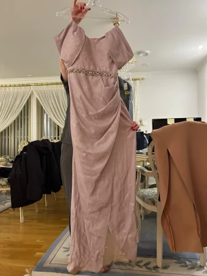 Ljusrosa glittrig långklänning - Elegant ljusrosa långklänning med glittrigt tyg och dekorativt pärlbälte i midjan. Klänningen har korta ärmar med voluminös design och en slits framtill. Perfekt för speciella tillfällen när du vill sticka ut.