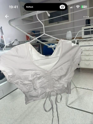 Vit croppad topp med snörning - Säljer en vit croppad topp med korta ärmar och snygg snörning som går att dra ihop mer mellan brösten, som ger en cool rynkad effekt. Toppen har v-ringning och är perfekt för dig som gillar trendiga detaljer och enkel stil.