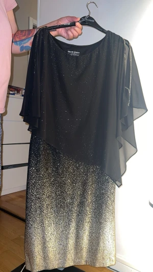 Svart och guld glittrig långklänning - Snygg långklänning i svart och guld med glittrigt tyg. Klänningen har ett lager av svart, transparent tyg upptill och skimrande guld nedtill. Perfekt för dig som vill sticka ut med en elegant look. Köpte den för ungefär 3500 kr och använts bara 1 gång