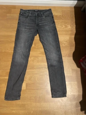 Grå raka jeans från Jack & Jones - Säljer ett par grå jeans från Jack & Jones med rak passform och klassisk femficksdesign. Jeansen har diskreta slitningar och är tillverkade i mjukt denimtyg. Perfekta för en avslappnad och stilren look.