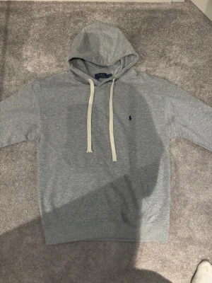Ralph Lauren hoodie - Snygg grå hoodie från Polo Ralph Lauren med vit dragsko och klassisk blå logga broderad på bröstet. Hoodien har känguruficka framtill och ribbade muddar vid ärmslut och nederkant. Perfekt för en chill och stilren look.