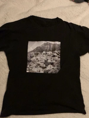 Svart t-shirt med fototryck - Svart t-shirt med ett stort svartvitt fototryck av ett bergslandskap och hus på bröstet. Klassisk rund hals och korta ärmar. Perfekt för dig som gillar stilrena plagg med coola tryck. Materialet känns mjukt och bekvämt mot huden.