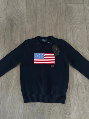 Mörkblå stickad tröja Polo Ralph Lauren - Mörkblå stickad tröja från Polo Ralph Lauren med amerikansk flagga i rött, vitt och blått på bröstet samt RL-broderi. Tröjan har rund halsringning och ribbade muddar vid ärmslut och nederkant. Perfekt för dig som gillar klassisk preppy stil.