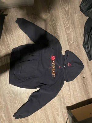 ABN University Hoodie - Säljer en svart hoodie med stor huva och känguruficka. På bröstet står det 'UNIVERSITY' i gult och rött med en cool grafisk detalj. På huvan finns även en liten röd brodyr. Perfekt för dig som gillar streetwear och vill ha en skön och stilren look.