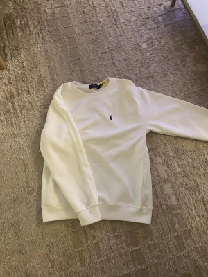 Vit sweatshirt från Polo Ralph Lauren - Klassisk vit sweatshirt från Polo Ralph Lauren med rund halsringning och den ikoniska lilla broderade loggan på bröstet. Tröjan har ribbade muddar vid ärmslut och nederkant. Perfekt för en clean och stilren look.