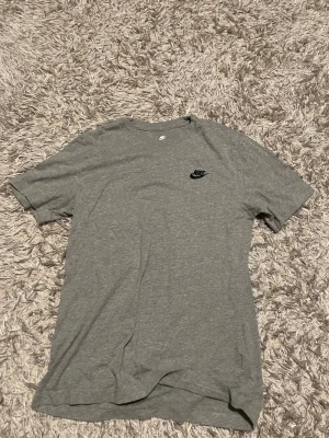Grå Nike t-shirt i bomull - Snygg grå t-shirt från Nike med klassisk logga på bröstet. Tillverkad i mjuk bomull och har en normal passform med korta ärmar. Perfekt till vardags för en sportig och enkel stil.