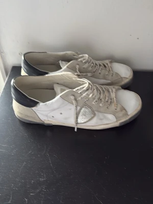 Vita Philippe Model sneakers med grå detaljer - Snygga vita sneakers från Philippe Model med grå och silverfärgade detaljer samt svart hälparti. Skorna har text på skosnörena och en unik paneldesign i mocka och läder. Perfekta för dig som gillar streetstyle och vill ha något som sticker ut. Defekter på snörena 