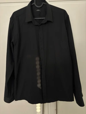 Svart skjorta från Tudor - Klassisk svart skjorta från Tudor med lång ärm och knappar framtill. Skjortan har en stilren design med vanlig krage och är tillverkad i ett mjukt tyg som känns bekvämt mot huden. Perfekt för dig som gillar en clean och enkel look.