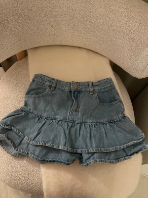 Blå volangskort i denim - Supersöt blå jeansskort med två volanglager och klassiska fickor fram. Gjord i mjukt denimtyg med knapp och dragkedja i midjan. 