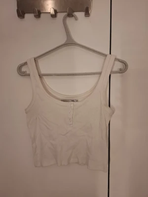 Vit ribbad linnetopp med knappar - Vit croppad linnetopp från lager 157 i ribbad bomull med knappar framtill. Toppen är ärmlös och har breda axelband, perfekt för en avslappnad och trendig look.