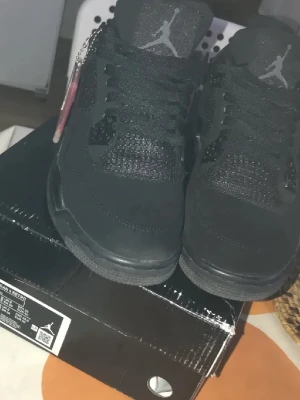 Svarta Air Jordan 4 Retro sneakers - Snygga helsvarta Air Jordan 4 Retro sneakers med ikoniska Jumpman-loggan på plösen och hälen. Skorna har en robust sula, meshdetaljer på sidorna och ovandel i mocka och textil. Perfekta för dig som gillar streetwear och klassisk sneakerstil.