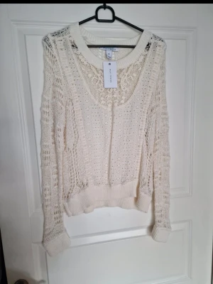Vit virkad stickad tröja Nelly - Trendig vit stickad tröja från Nelly med virkat mönster och transparenta partier. Tröjan har rund halsringning, långa ärmar och ribbade muddar vid ärmslut och nederkant. Perfekt för lager på lager och ger en cool, avslappnad look.