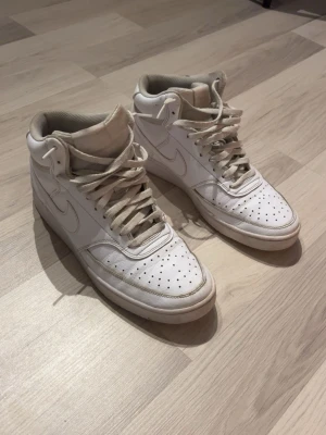  Nike high skor - Säljer ett par klassiska vita high top sneakers från Nike. Skorna har perforerad tå, vita snören och diskret Nike-logga på sidan. Tillverkade i läder med gummisula för bra grepp. Perfekta för dig som gillar retrostil och streetwear.