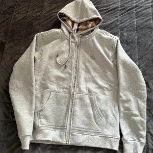 Burberry Tröja - En läcker grå Zip Hoodie i märket BURBERRY, vacker elegant tröja som verkligen sticker ut i mängden. Märket Burberry kommer ursprungligen från London och är idag ett klädesmärke som förmedlar stiliga lyx kläder.  Tröjans storlek är M