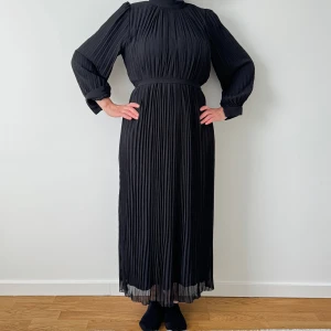 Klänning - Oanvänd plisserad klänning i svart med eller utan knytskärp. Storlek s/m upplevs som onesize. Knappar vid handled och nacke.