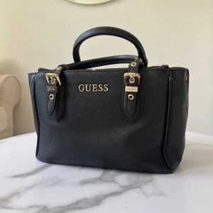 Guess handväska - Jättefin!! De guldiga på handtagen har rostat lite annars superfin🥰 Det finns även ett längre band som kommer med. Längd: 24 cm Höjd: 16 cm