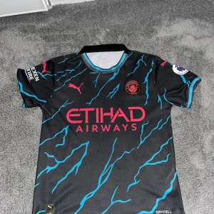 Man City Tredjeställ - Uppgradera din fotbollsstil med Manchester Citys tredjeställ 23/24.Tillverkad av andningsbart material för maximal komfort. Modern passform och iögonfallande design gör att du sticker ut både på och utanför planen. Perfekt för alla fotbollsälskare!