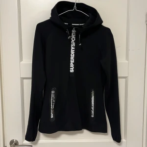 Superdry Sport hoodie - Väldigt fin hoodie från Superdry Sport. Både sportig och snygg till vardagligt bruk. I bomullsmaterial, eller samma material som vanliga hoodies. Fräcka detaöjer med tryckt text på blixtlåsen och bak nertill. Storlek S, i fint skick!!