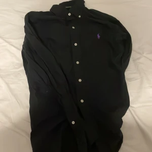 Svart polo ralph lauren skjorta - Svart med lilla logo, slim fit men passar olika storlekar; perfekt för festiva aktiviteter. Prisen kan diskuteras.