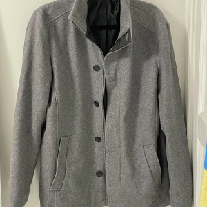 Jack & Jones vinterkappa grå ullblandning - Grå jacka från jack and jones med ståkrage, två fickor, dragkedja, och knappar. Nästan nytt skick, inga skönhetsfel vad jag kunde hitta. Väl omhändetagen!