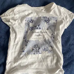 Vit T-shirt med blommor på. Använd ungefär 10 gånger, inga skador förutom att det står ”saga” på baksidan längst upp, men inget man ser när tröjan är på.