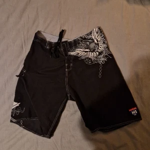 Surfshorts m. Rocktryck - Svarta surfshorts med häftigt tryck. De har kardborre och snöre i midjan + en sidficka, köpta secondhand men aldrig använda!