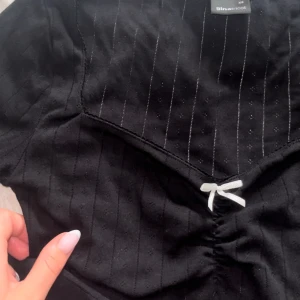 Svart topp - Svart topp med lite håll i. Ribbad aktig topp med vit rosett. Fin urringning med scrunch i fram. 