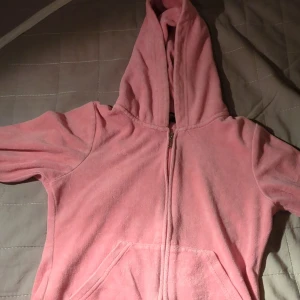 Rosa fleecejacka från Juicy Couture - En mjuk och bekväm rosa fleecejacka från Juicy Couture. Jackan har en dragkedja framtill och en huva. Den har också två framfickor.